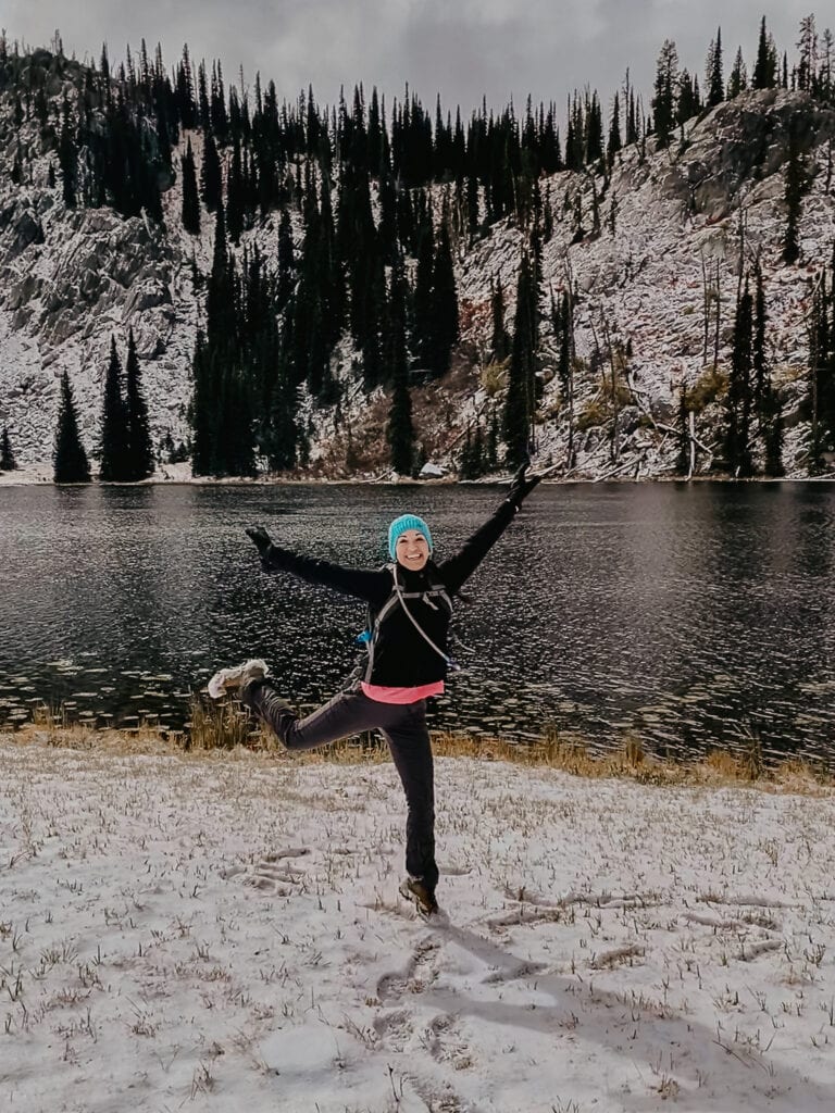 Woman hiker jumps in the air in delight in front of a snowy lake.