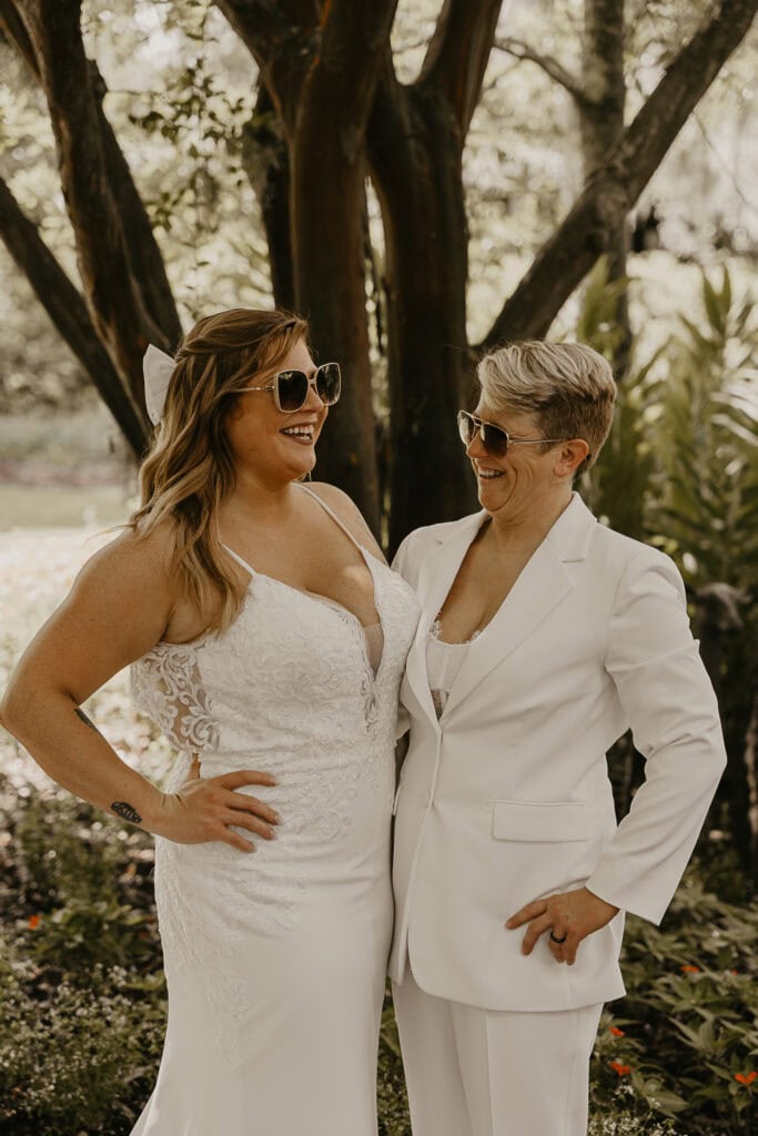 An LGTBQ+ couple laughs together outside on their wedding day while wearing sunglasses.