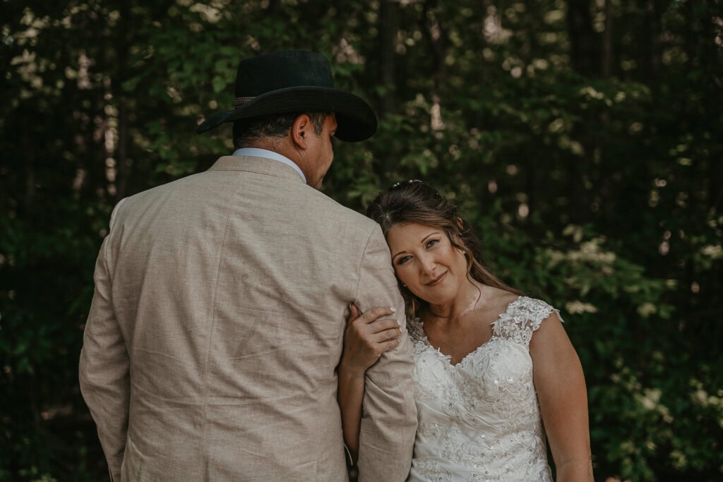 Anita looks at the camera while holding John's arm and resting her head on this shoulder as he faces the opposite direction.