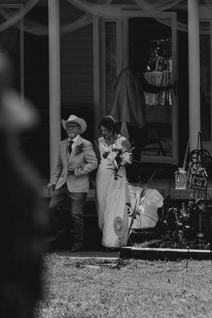 A bride and her dad walk arm in arm down the front steps of her home.