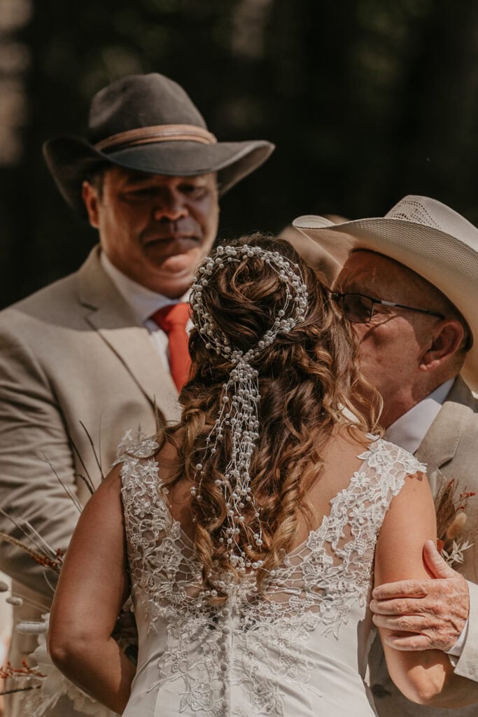 The father of the bride gives her a kiss while the groom smiles at her.