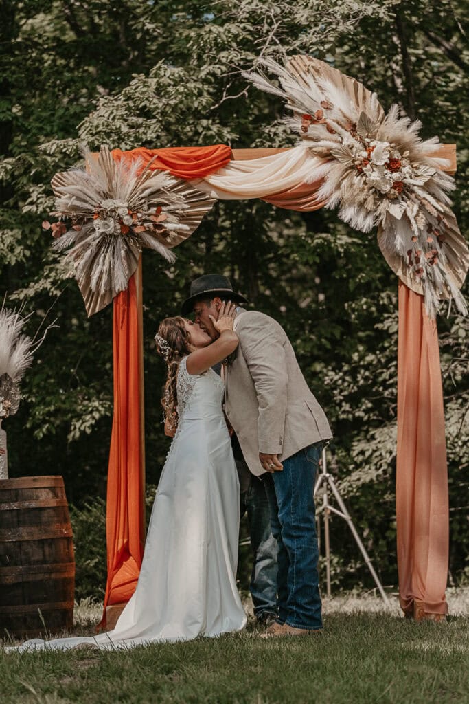 A couple shares a first kiss during their wedding, under a wooden arch that's lined with flowy scarves.