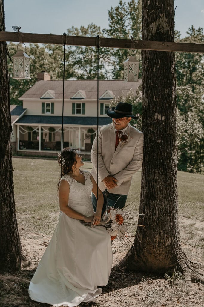 Anita sits on the tree swing that John built, as he looks at her, leaning on the tree. They're in their wedding atiire.