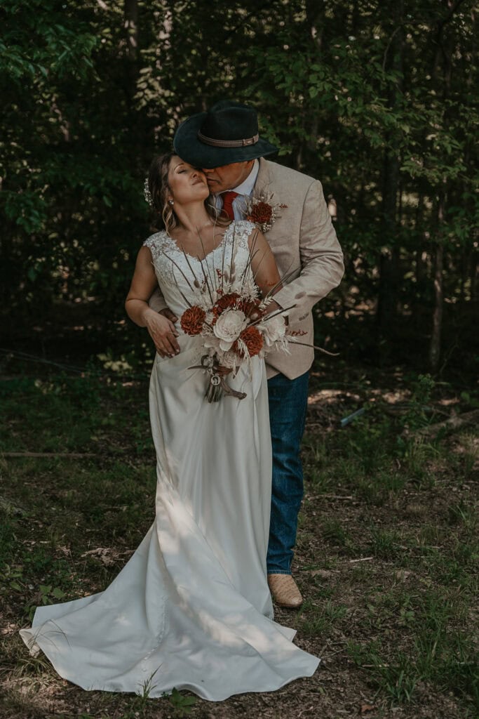 A groom kisses the bride as he stands behind her with his arms wrapped around her waist.