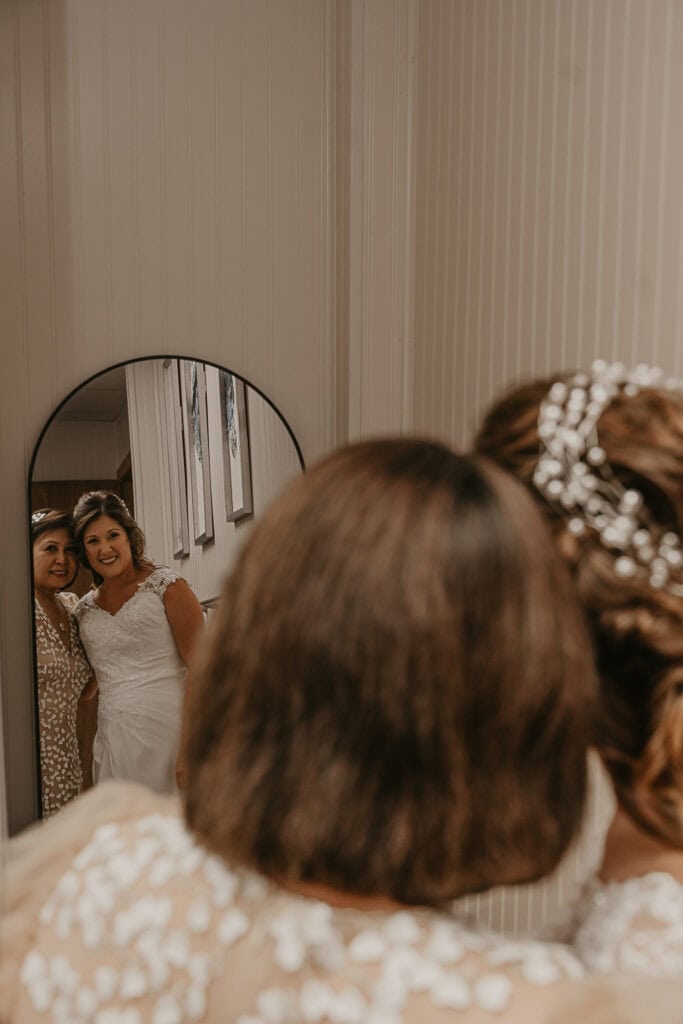 A bride and her aunt side hug and look at each other in the mirror before the wedding ceremony.