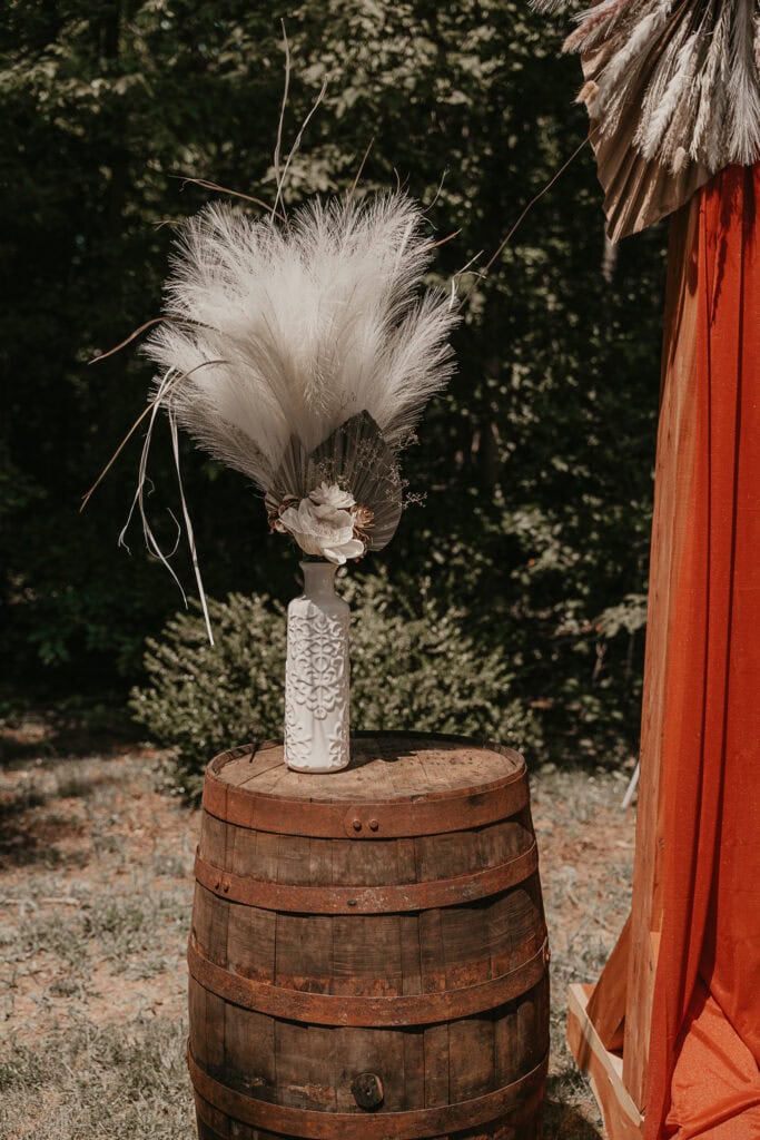 Decorations at the front of the wedding area of an outdoor wedding include a wooden barrel and a neutral bouquet in a vase.