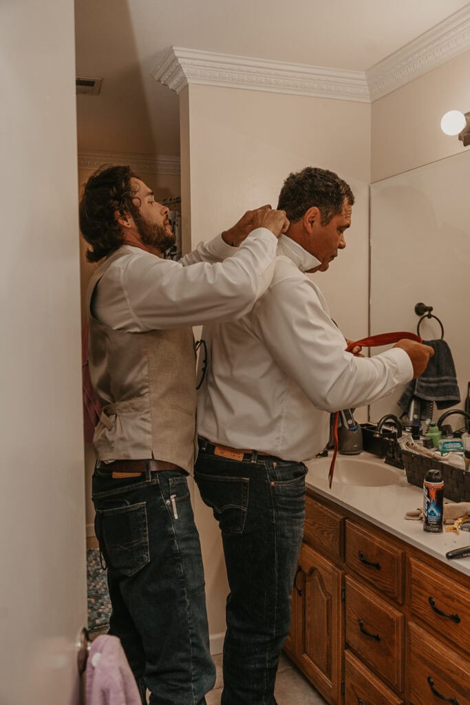 The groom's son helps him get ready by adjusting his tie under the collar.