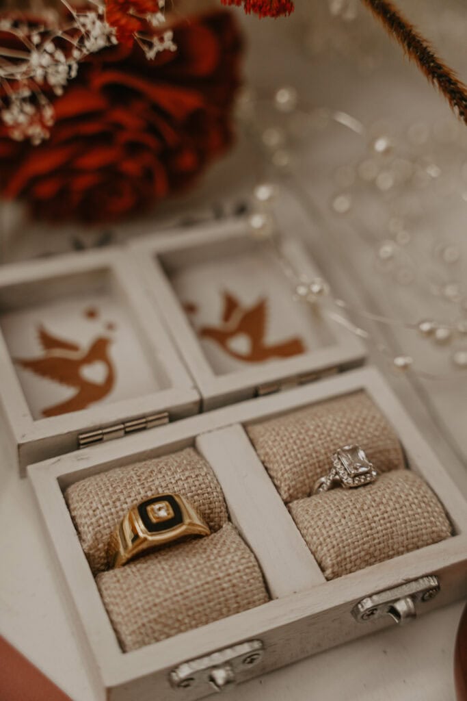 A closeup of bride and groom's rings in a wooden box with the bouquet in the background.