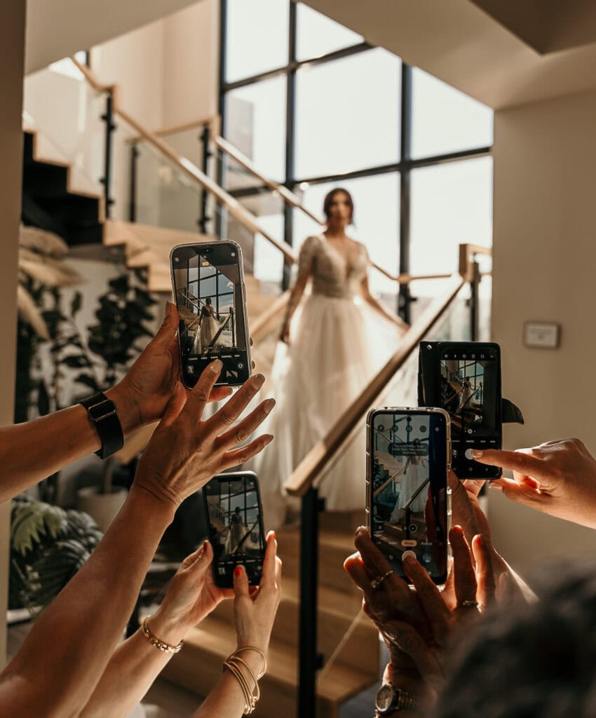 Amaris' friends take photos of her on their phones as she walks down the stairs in her wedding gown.