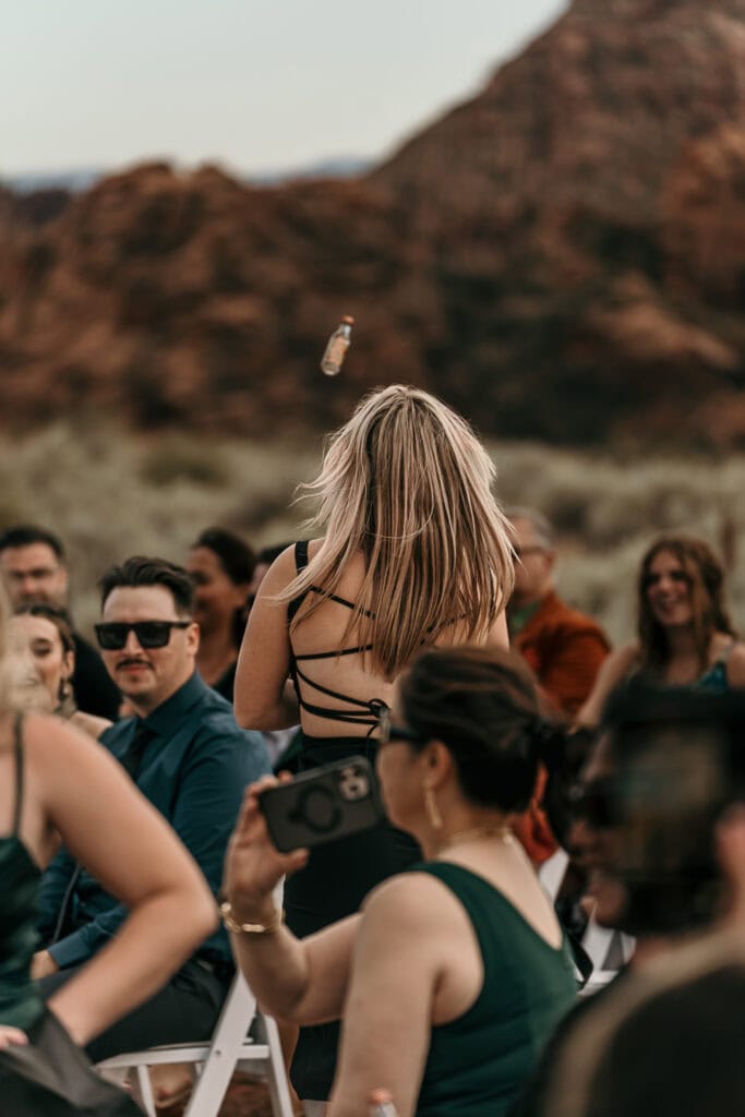 A shot girl tosses a bottle of liquor to guests at an outdoor wedding