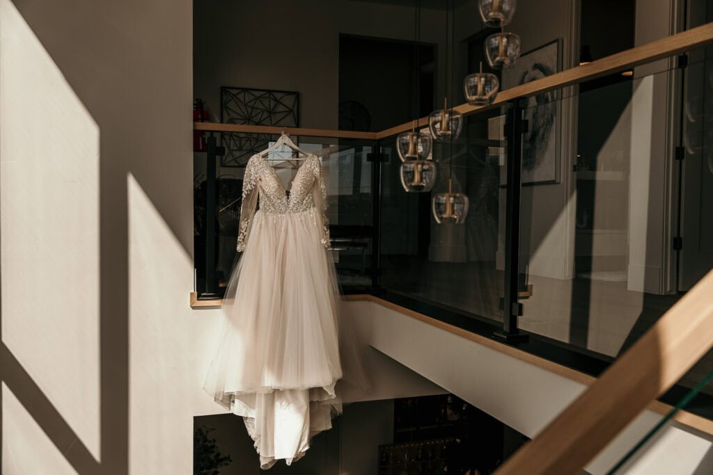 A wedding gown hangs in a stairwell as sunlight shines on it.