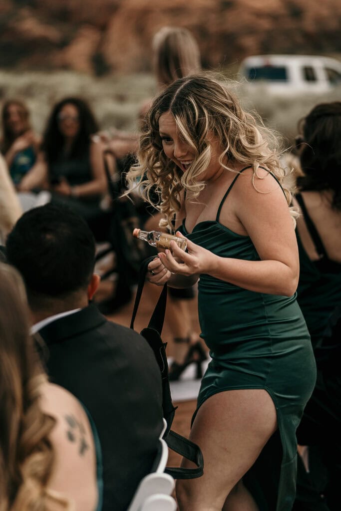 A shot girl hands a bottle of alcohol to a guest at an outdoor wedding.