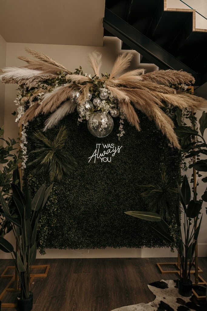 A photobooth area at a reception made up of a disco ball, greenery, and a neon 