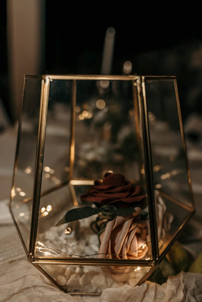 A fake rose sits in a glass and gold holder with twinkle lights.