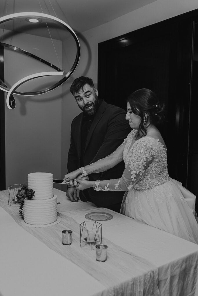 A bride pretends its really hard to cut the cake while the groom looks at her laughing.