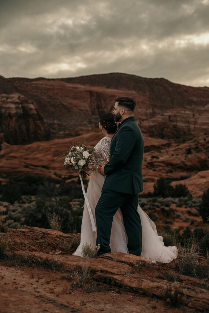 Bride and groom look out over the red rocks in Utah as the sun sets behind them.