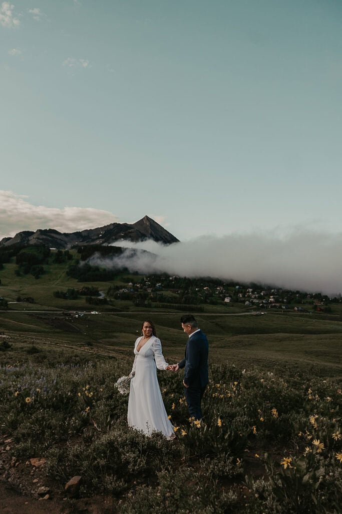 dreamy crested butte elopement couple in a field of wildflowers with houses covered in smoke in the backdrop