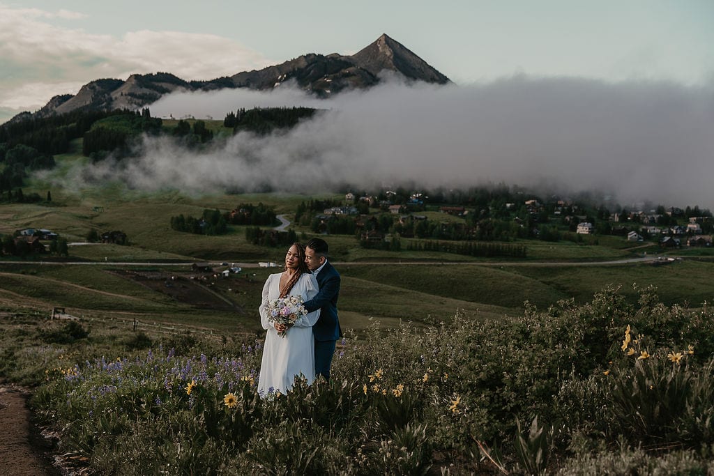 dreamy crested butte elopement couple in a field of wildflowers with houses covered in smoke in the backdrop