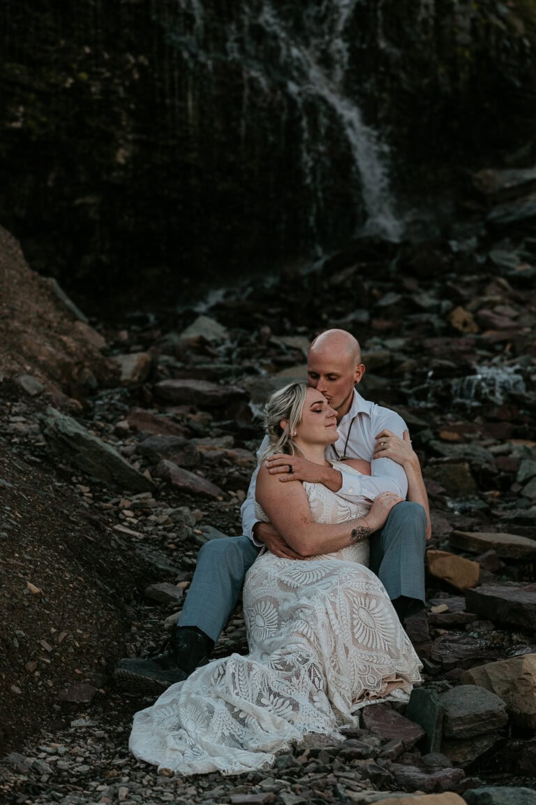 A couple snuggles while sitting in front of a waterfall. They're in elopement attire.