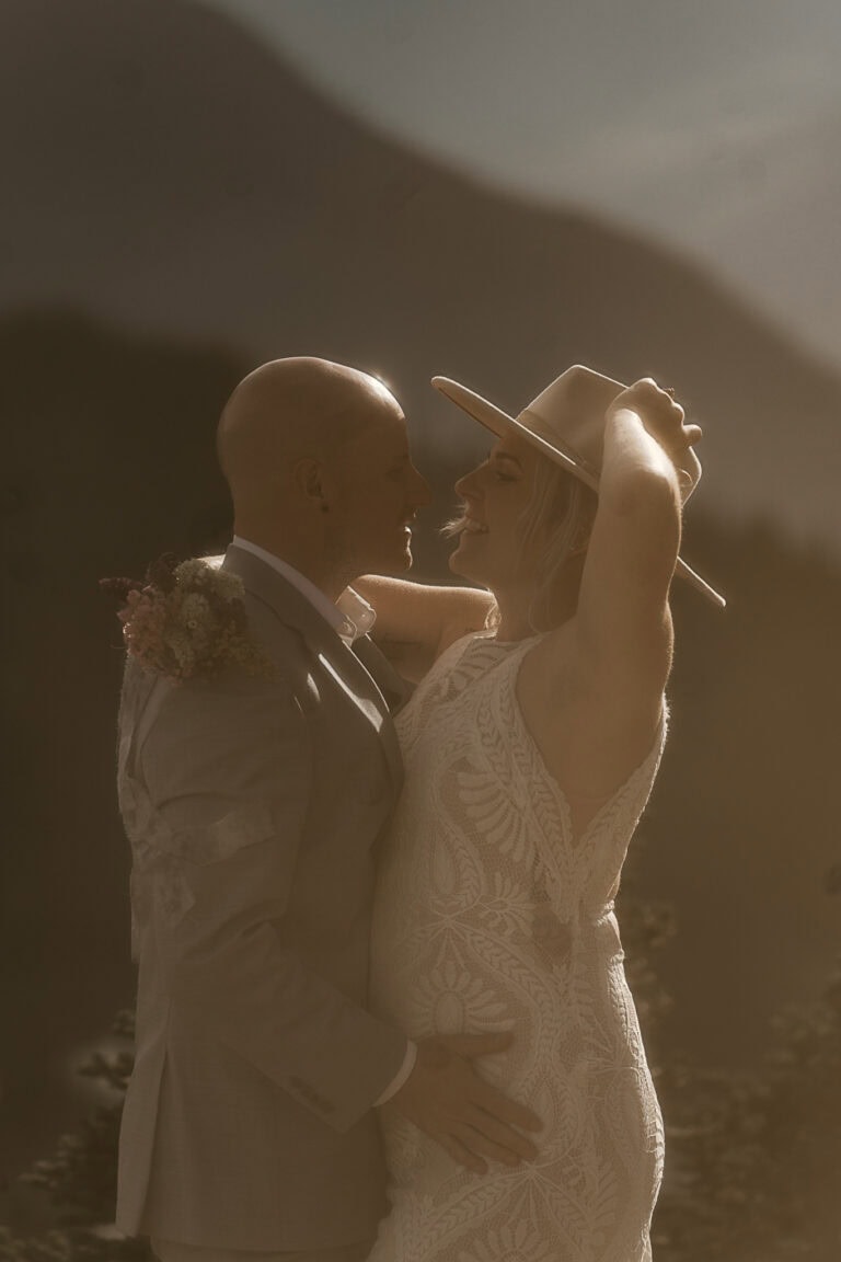 A bride holds onto her boho hat while her and the groom embrace outside.