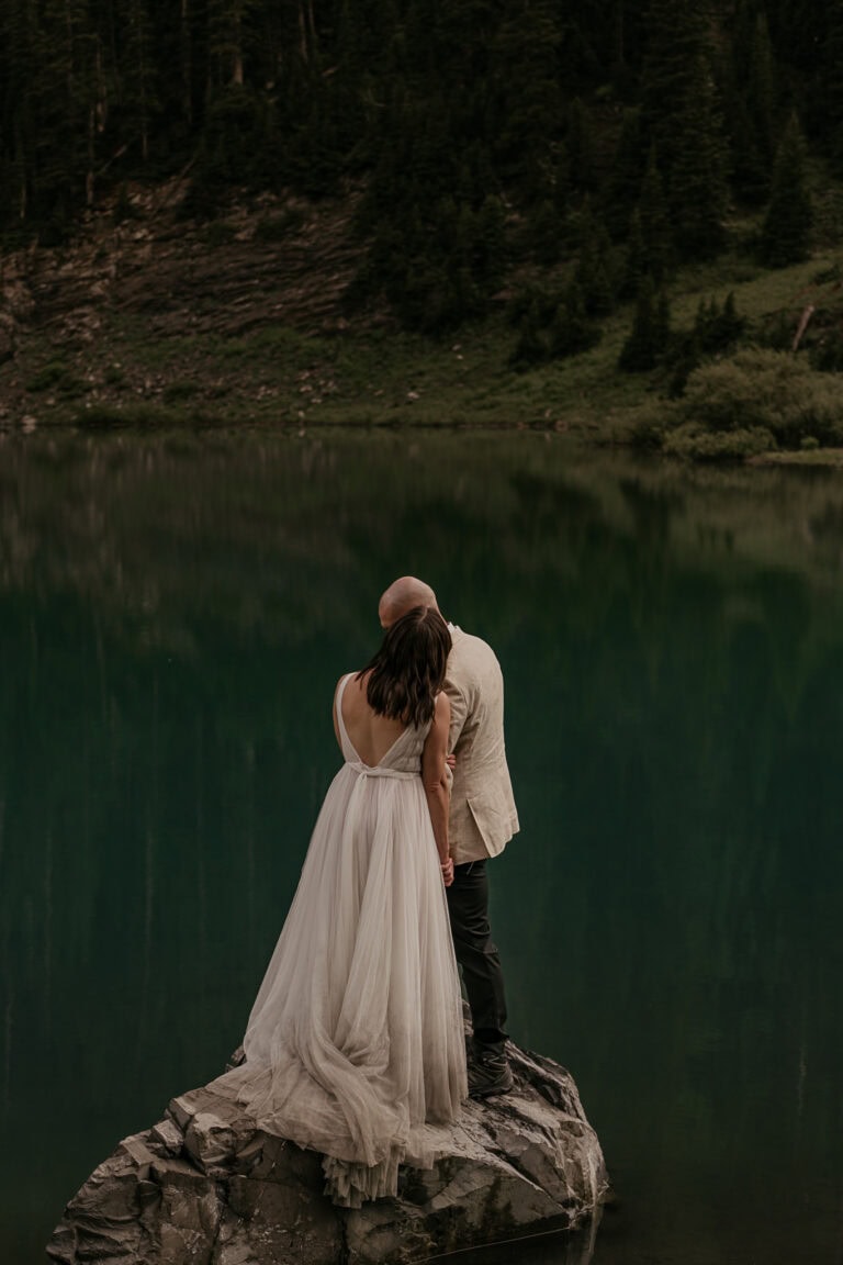 A bride and groom stand on a rock looking out over an alpine lake.