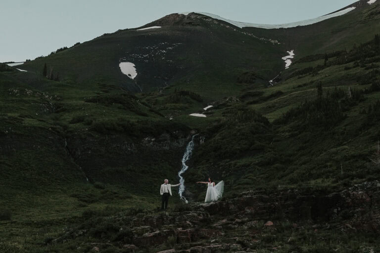 A bride and groom stand on each side of a small cascade in the mountains and reach towards each other.