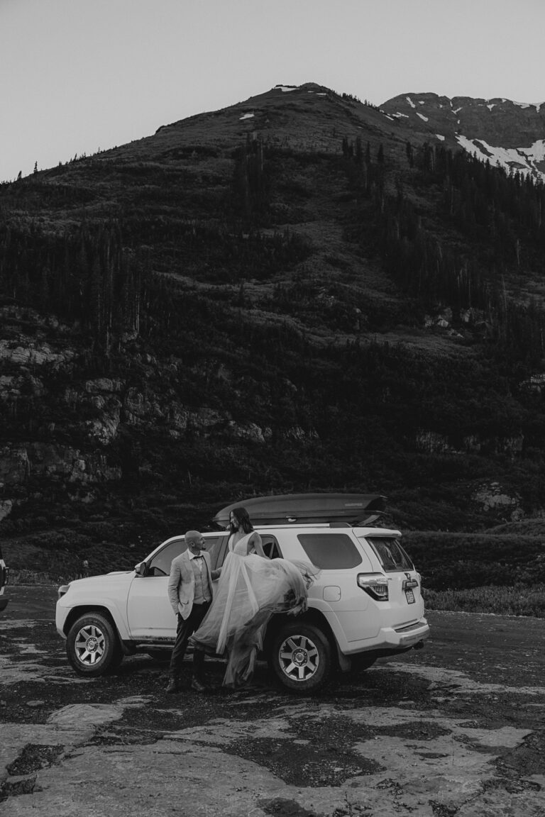 A black and white photo of a bride and groom standing on the running board of their Forerunner in front of the mountains.