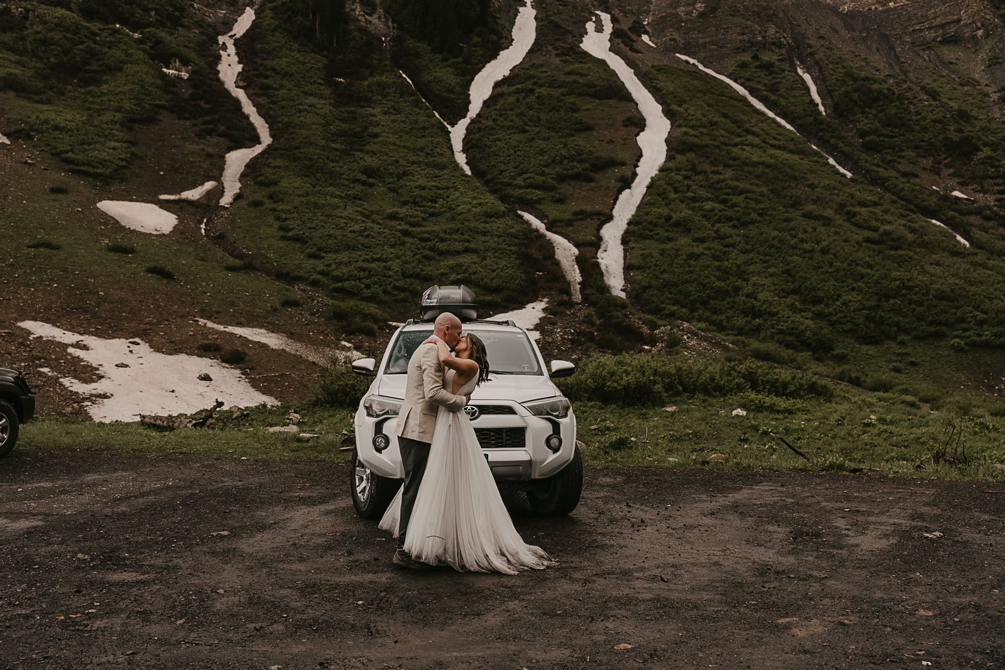 A bride and groom kiss in front of their SUV and snowy mountains.