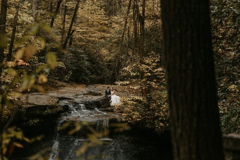 A couple sits on top of a waterfall in autumn. They're in wedding attire.
