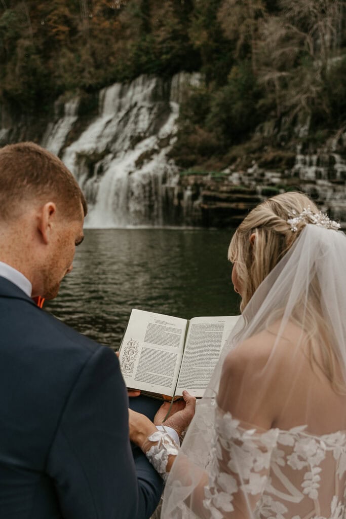 A couple in wedding attire sit and read together. A waterfall is in the background.