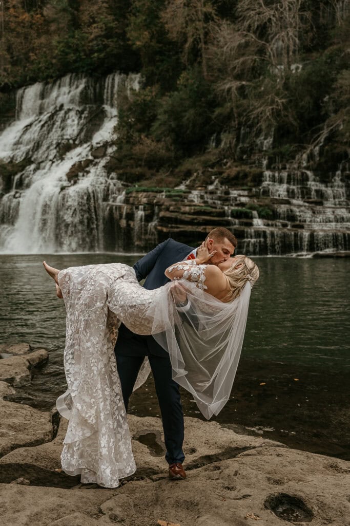 A groom dip kisses his bridge while they stand in front of a waterfall.