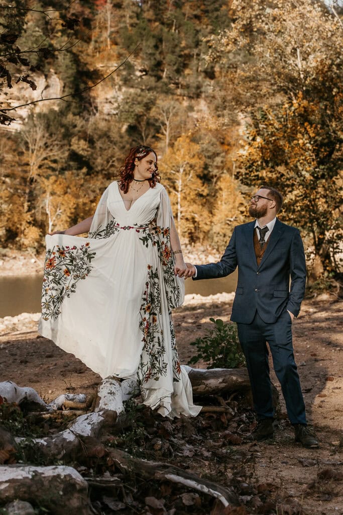 A bride playfully twirls her dress while standing on a large tree root as her husband looks on. They're holding hands.