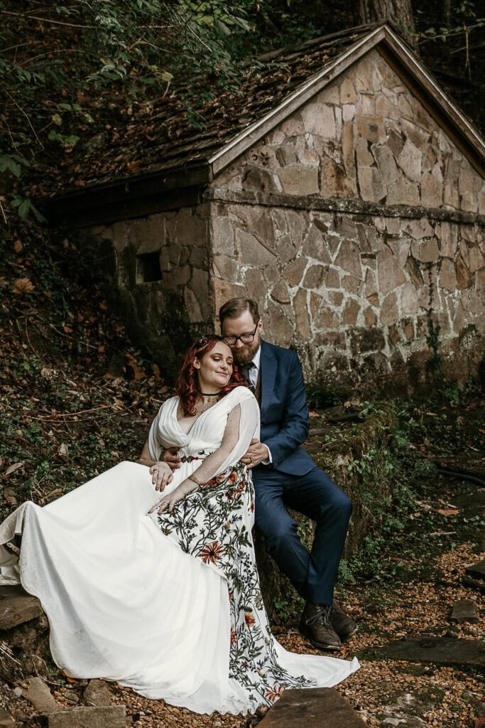 A couple sits closely on a rock ledge with a stone building behind them. Their eyes are closed.