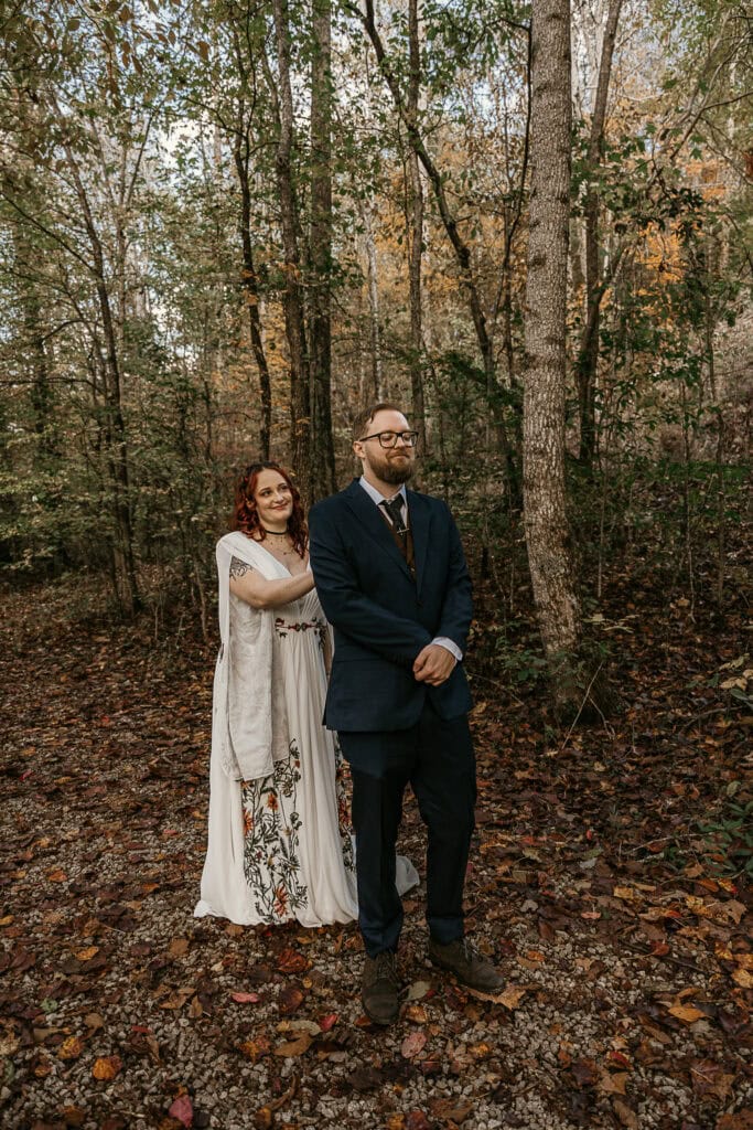 A woman stands behind a man on a trail. They're in wedding attire. He faces the same direction as her.