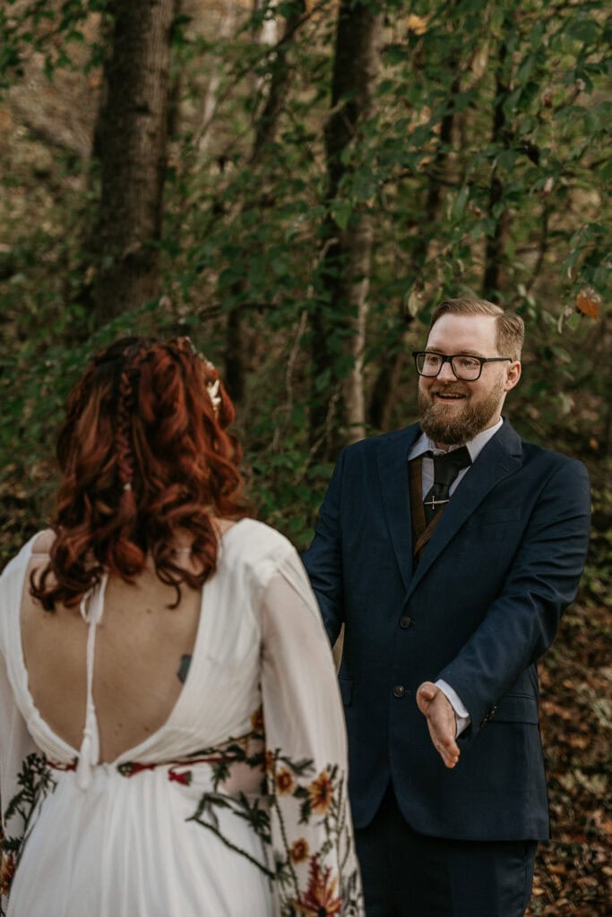 A man is beaming at the sight of his bride to be while they stand outside in the woods.