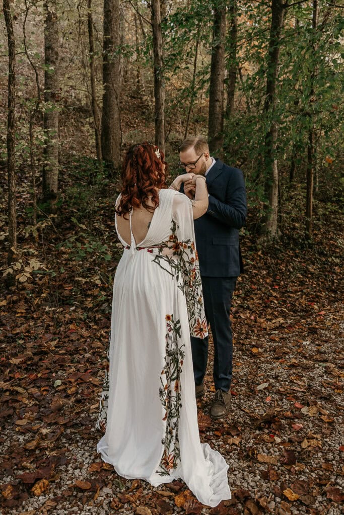 A couple in wedding attire stand on a trail in the forest facing each other. He kisses her hand.