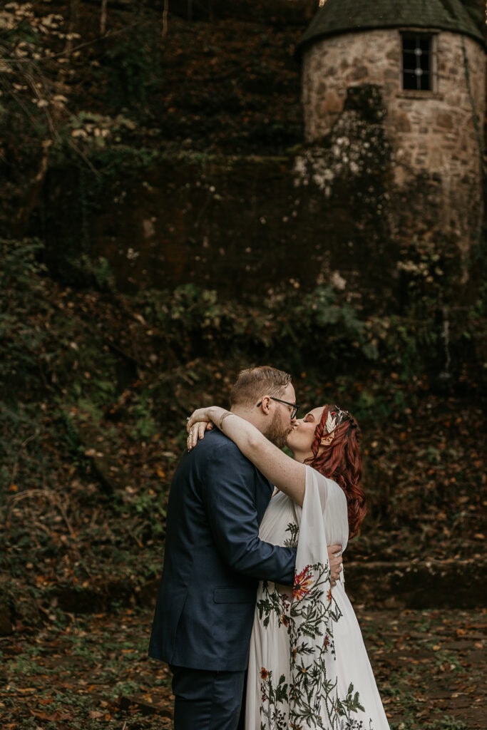 A couple kisses in front of a stone turret. They're in wedding attire.