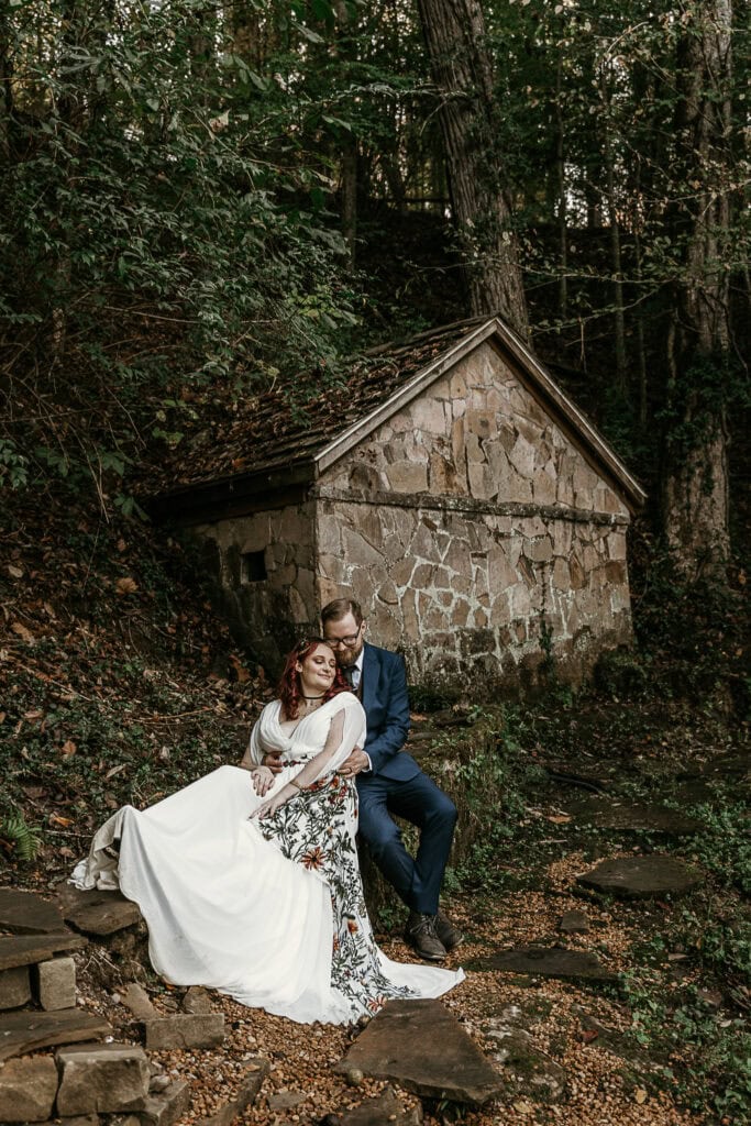 A couple shares a cozy moment while sitting on a rock wall in front of an old stone building. They are in wedding attire.