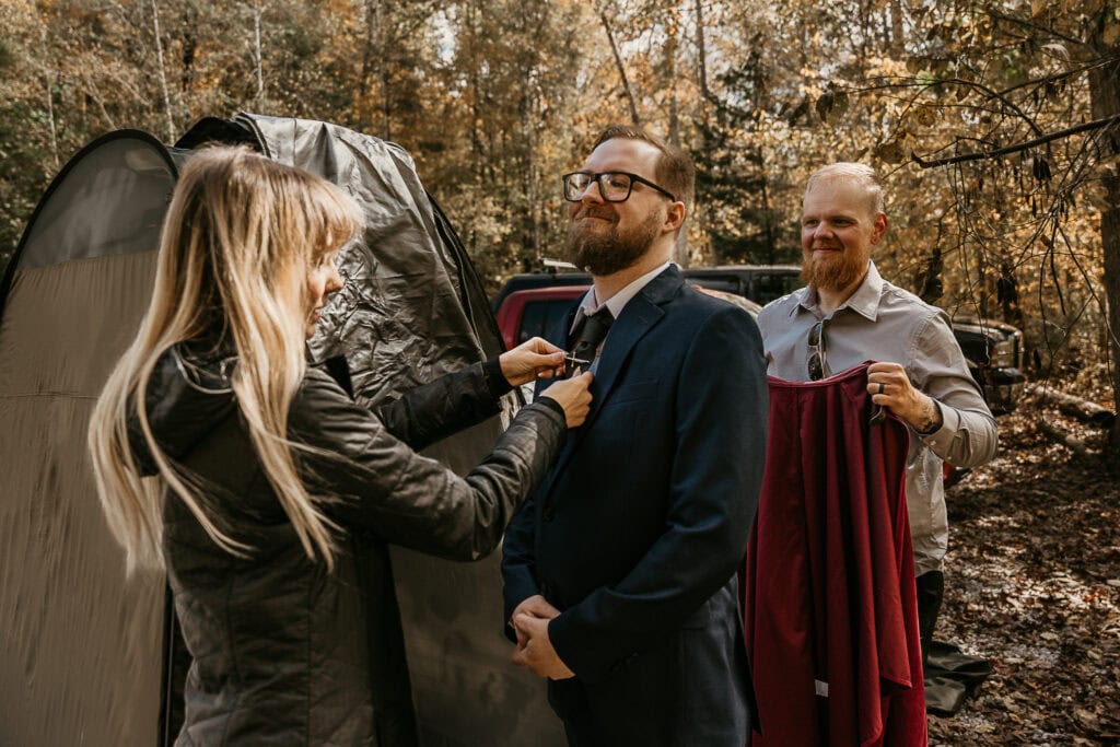 Friends help a groom get ready outside in a parking lot. There is a changing tent next to them.