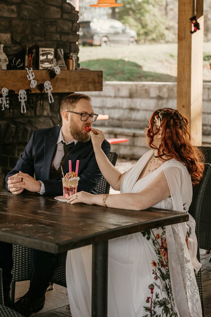 A bride feeds the groom a bite of a strawberry from their shared milkshake.
