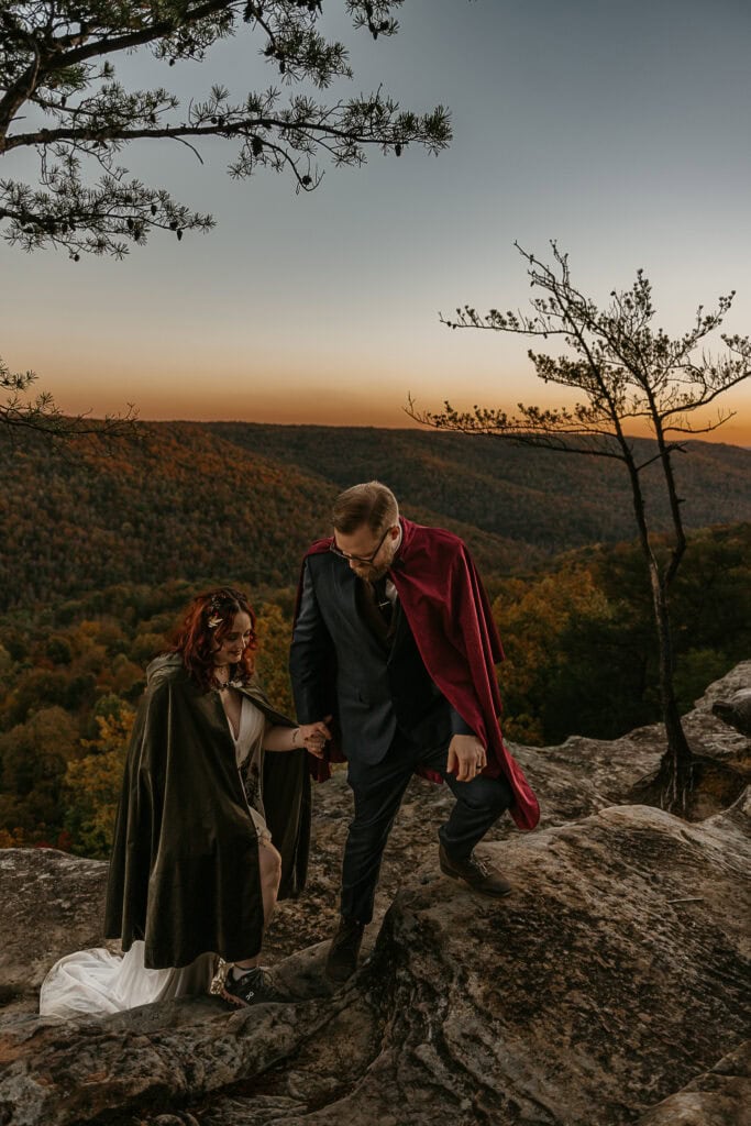 A couple in wedding attire and capes climb up rocks while holding hands. Fall colors and an overlook are behind them.