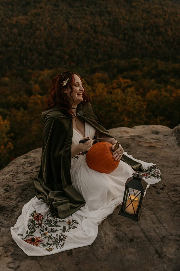 A woman in wedding attire sits on a cliff's edge carving a pumpkin while laughing.