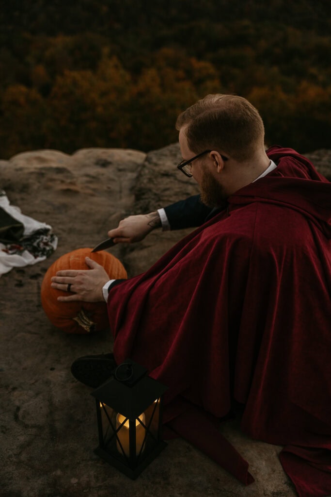 A man in a cape carves a pumpkin while sitting on the edge of an overlook.