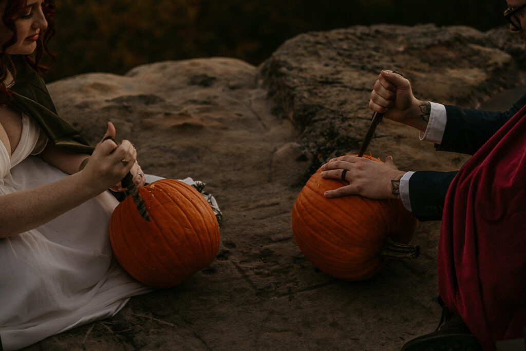 A couple carves pumpkins together during their elopement. They are on a cliff.