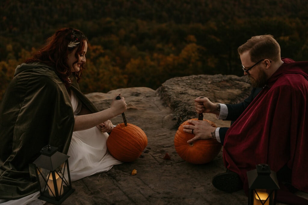 A couple laughs while carving pumpkins together as the sun sets. They are sitting on an overlook with lanterns.