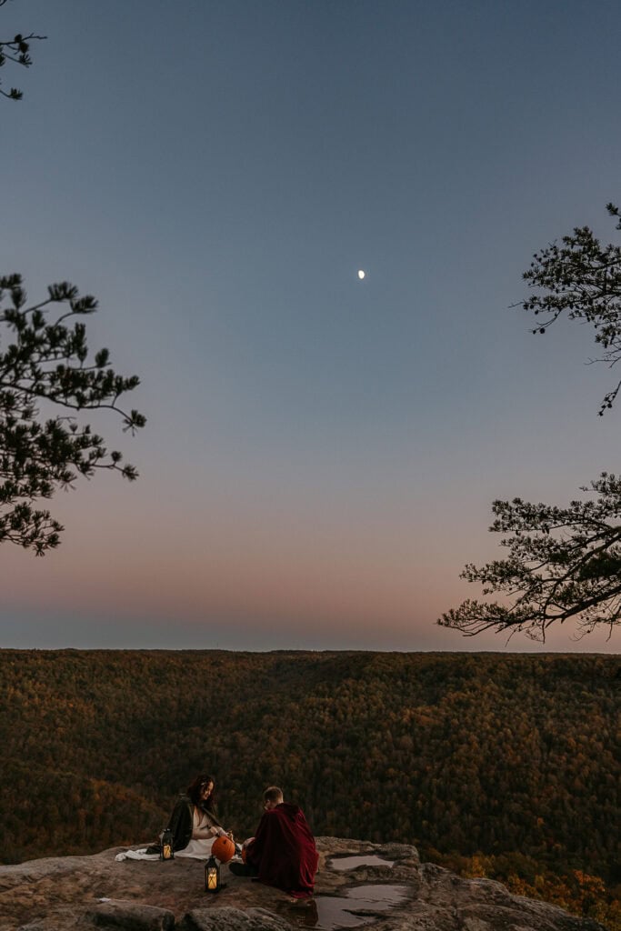 The moon shines overhead as a couple in wedding attire sit on an overlook carving pumpkins with lanterns.