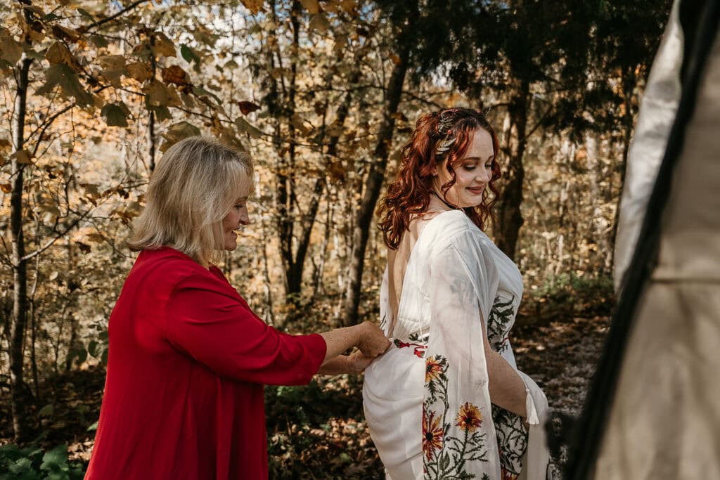 Mom zips up the back of the bride's dress. They are outdoors.