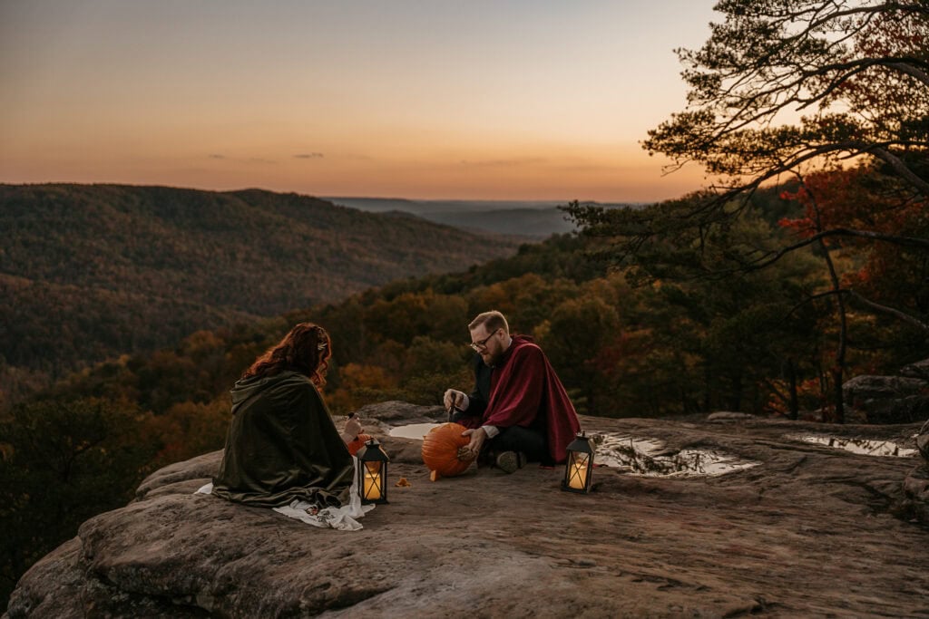 A couple concentrates on carving pumpkins as the sun sets around them. They are on an overlook and fall colors are in the valley.