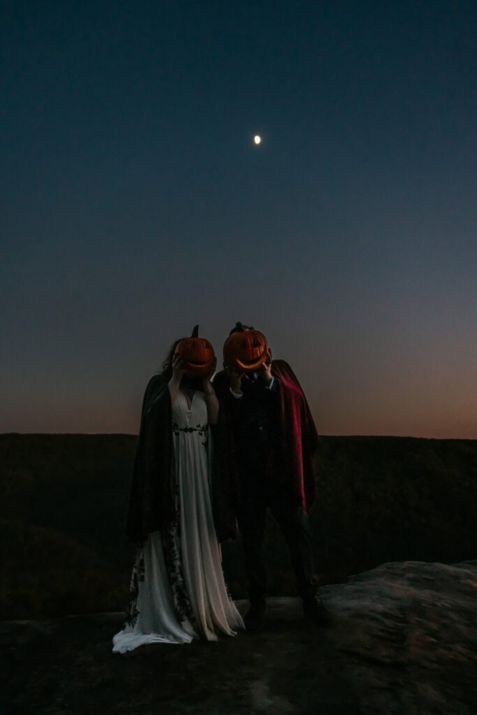 A couple in wedding attire hold carved pumpkins in front of their faces while standing under the moon on a rock overlook.