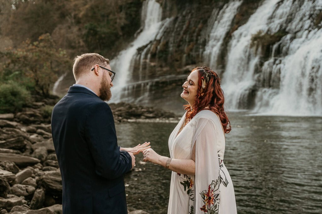 A couple looks at each other happily during their ring exchange. Twin Falls is in the background.